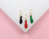 Three enamel figa hand charms in black, red, and green with gold bails rest on a white display against a pink background.
