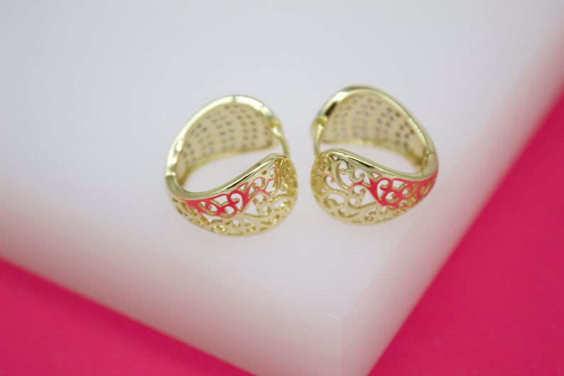 A pair of gold hoop earrings with intricate filigree designs rests on a white display surface against a vibrant pink background.