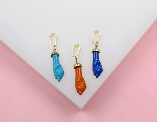 Three colorful fist-shaped enamel charm pendants in turquoise, orange, and blue hang against a white and pink background.