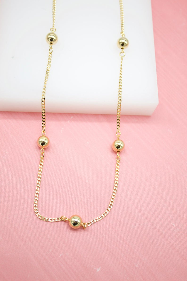 A delicate gold chain necklace features evenly spaced polished bead stations displayed against a pink background.