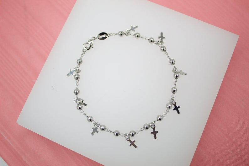 A silver bead charm bracelet featuring dangling mini cross charms rests on a white display stand against a pink background.