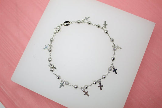 A silver bead charm bracelet featuring dangling mini cross charms rests on a white display stand against a pink background.