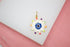 A gold evil eye charm featuring colorful enamel details rests on a white display surface.