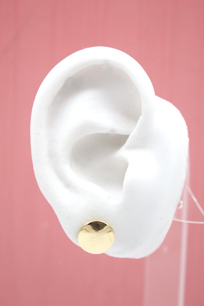 A white mannequin ear displays a polished gold disc stud earring against a pink background.