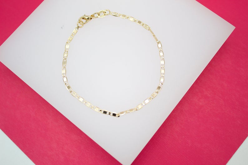 A delicate gold link bracelet rests on a white display stand against a vibrant pink background.