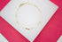 A delicate gold link bracelet rests on a white display stand against a vibrant pink background.