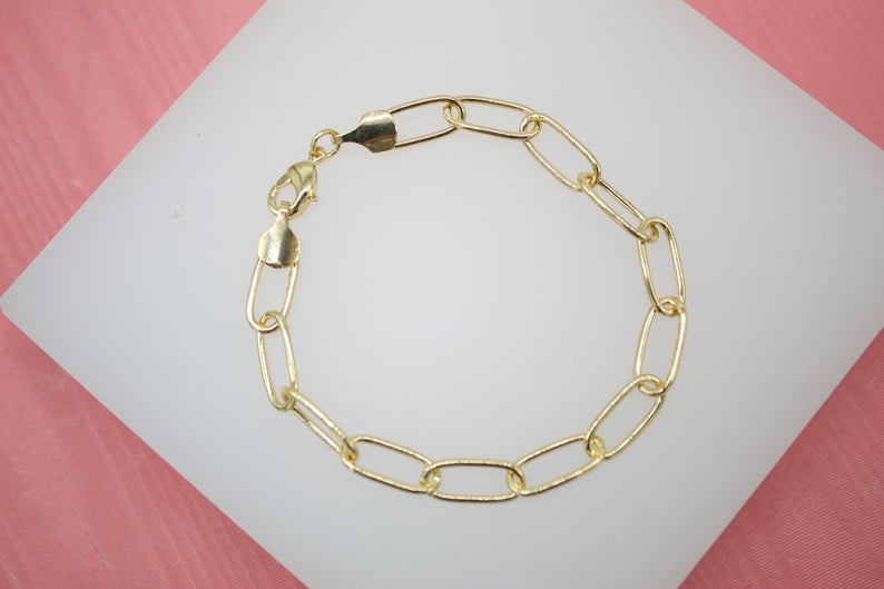 A gold paperclip chain bracelet rests on a white display surface against a pink background.
