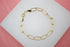 A gold paperclip chain bracelet rests on a white display surface against a pink background.