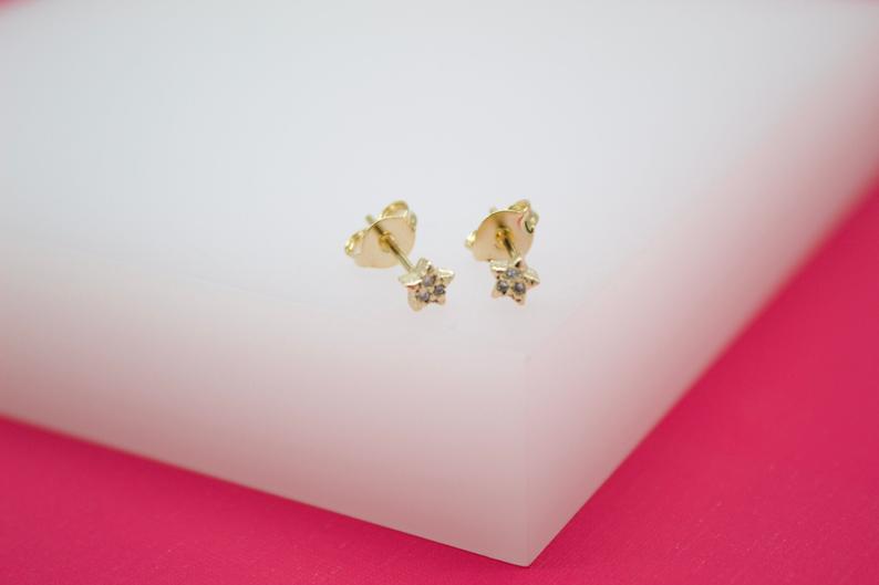 A pair of dainty gold star stud earrings rests on a white display surface with a pink border.