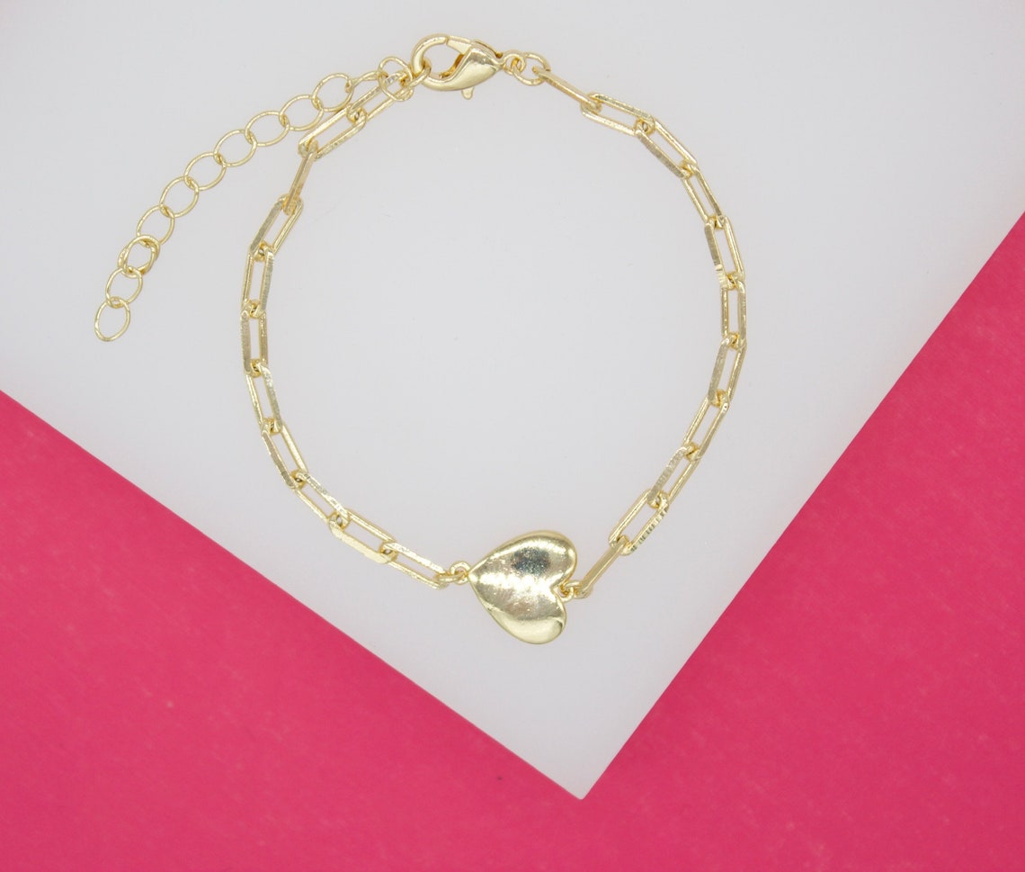 A delicate gold chain bracelet features an interlocking paperclip design and a polished heart charm, displayed against a dual-toned white and pink background.