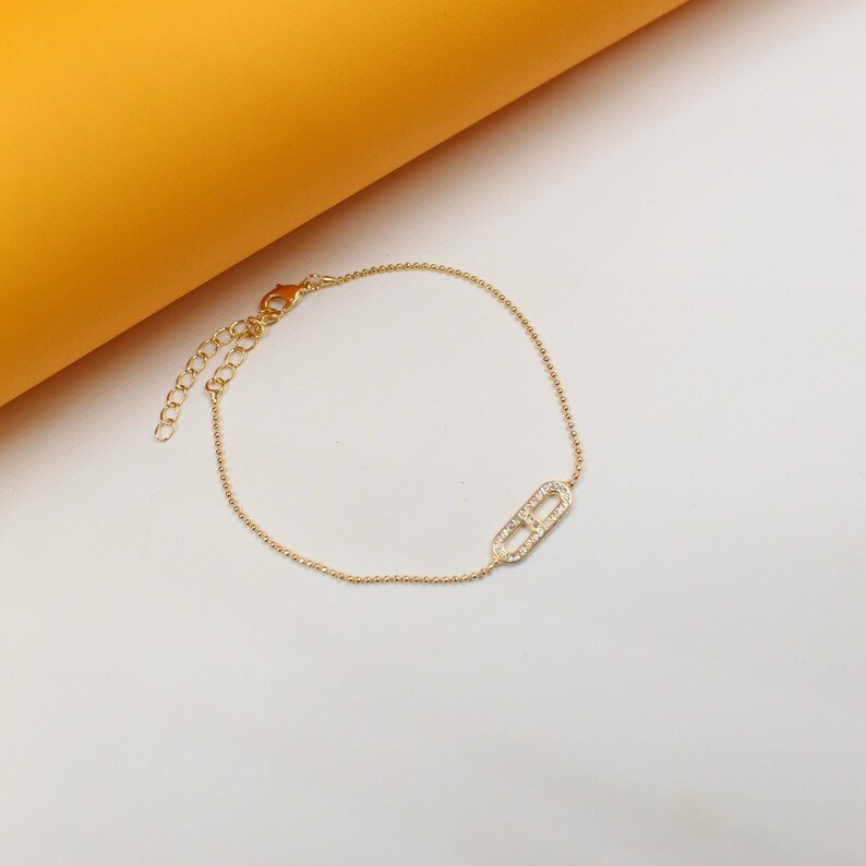 A delicate gold beaded bracelet features an interlocking geometric link encrusted with sparkling rhinestones, displayed against a white and orange background.