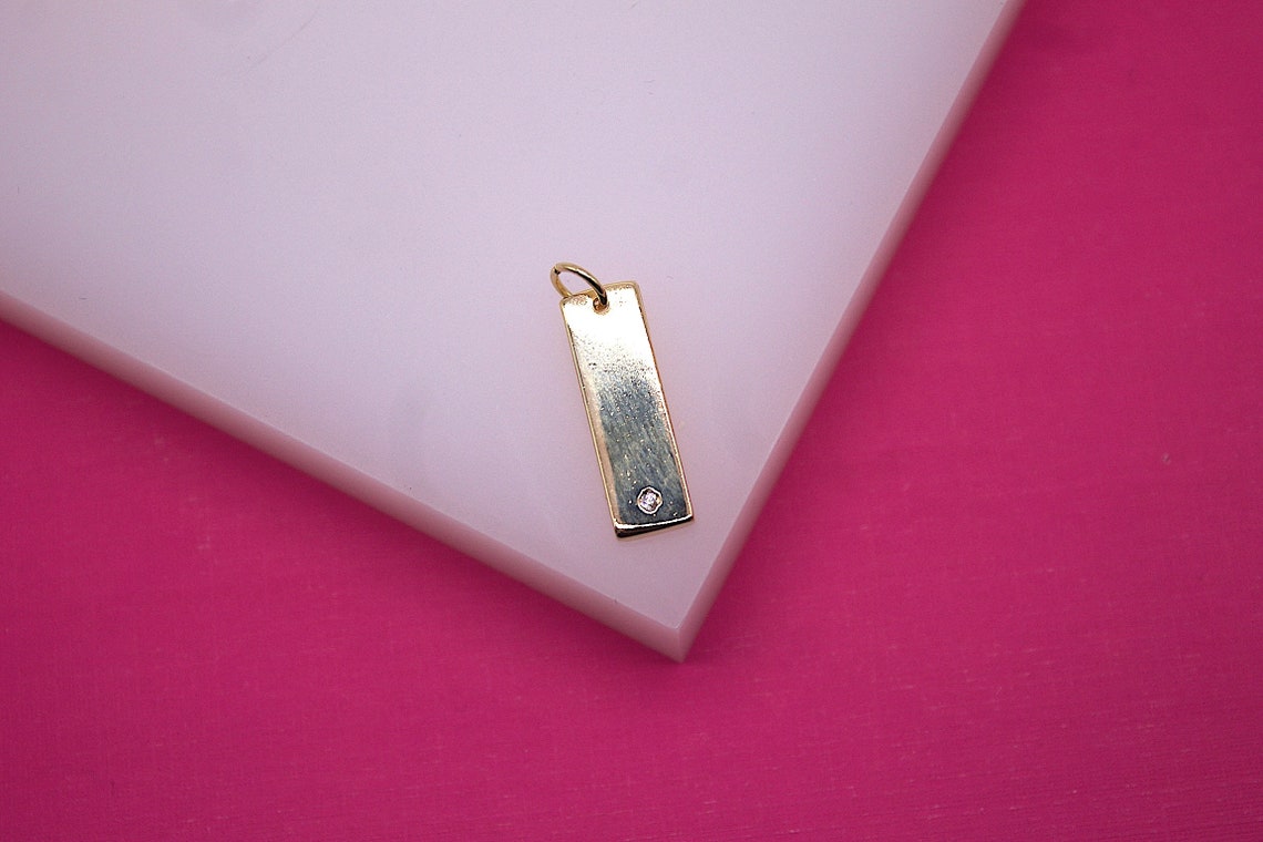 A delicate gold bar pendant featuring a sparkling diamond accent rests on a white display surface against a vibrant pink background.