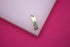 A delicate gold bar pendant featuring a sparkling diamond accent rests on a white display surface against a vibrant pink background.