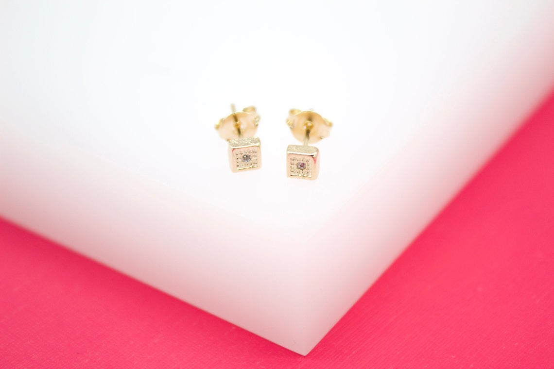 A pair of delicate gold square stud earrings with center stones rest on a white surface.