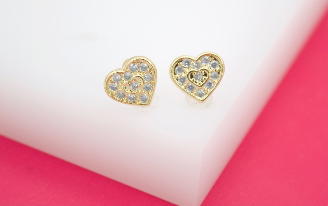 A pair of gold heart-shaped stud earrings featuring sparkling cubic zirconia stones rests on a white display block.