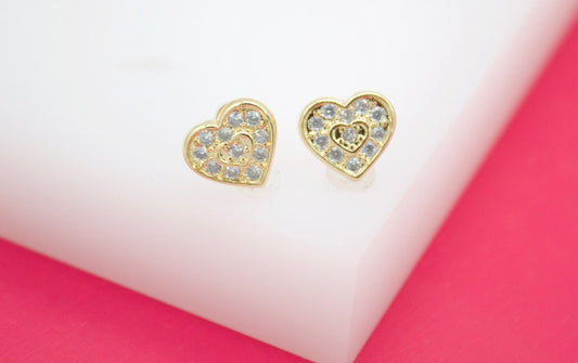 A pair of gold heart-shaped stud earrings featuring sparkling cubic zirconia stones rests on a white display block.