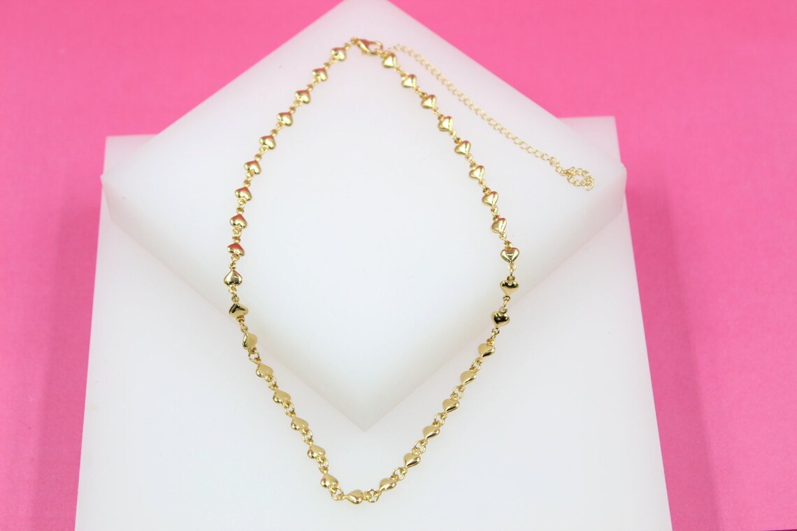 A delicate gold chain necklace featuring small heart charms rests on a white display block against a vibrant pink background.