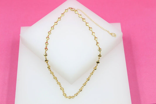 A delicate gold chain necklace featuring small heart charms rests on a white display block against a vibrant pink background.