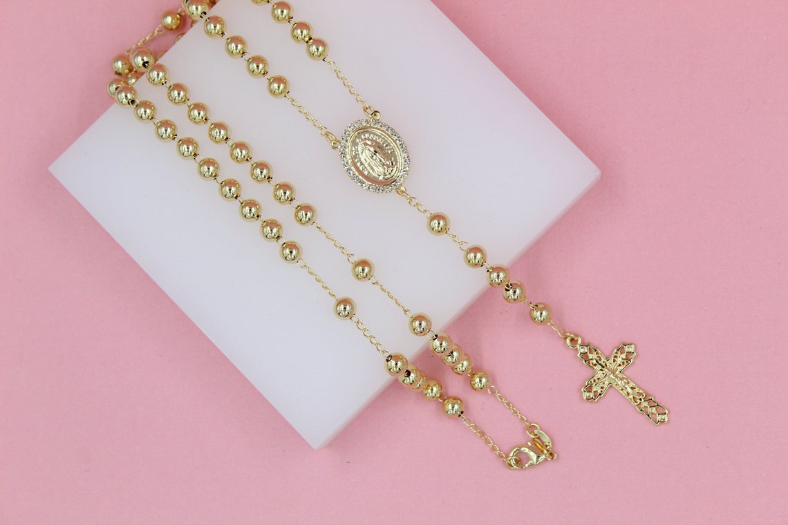 A delicate gold rosary necklace with polished spherical beads, a miraculous medal centerpiece, and an ornate crucifix rests on a white block against a soft pink background.