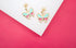 A pair of gold butterfly earrings featuring enamel wings in turquoise and pink rests on a white display against a vibrant pink background.