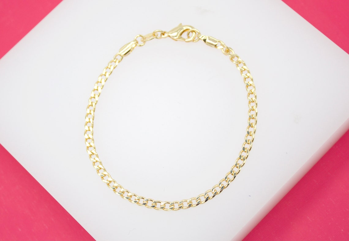 A delicate gold-plated chain bracelet rests on a white display surface against a vibrant pink background.