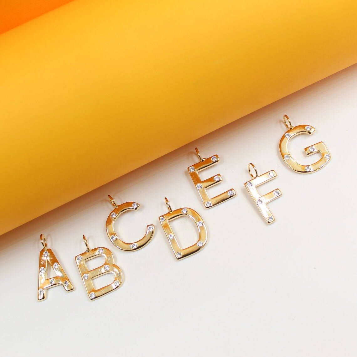 Sparkling gold initial letter pendants from A to G rest on a bright yellow and white background.