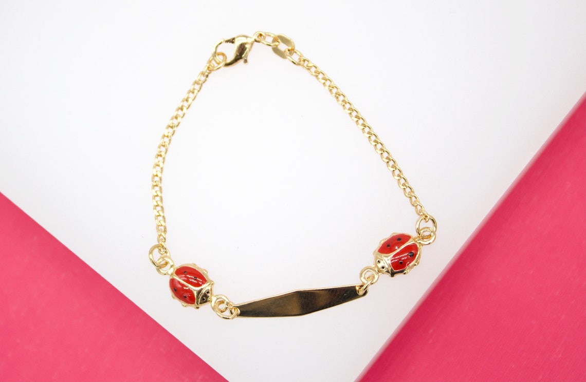 A gold-plated children's identity bracelet features two red enameled ladybug charms flanking a blank nameplate.