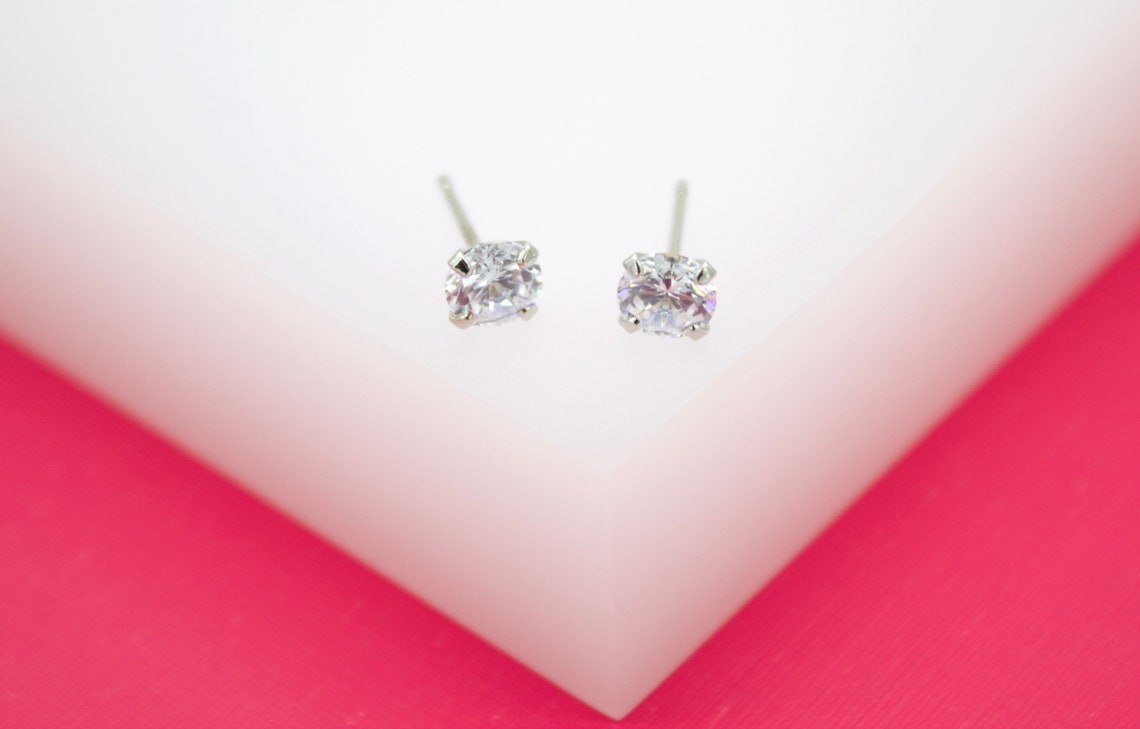 A pair of sparkling solitaire cubic zirconia stud earrings rests on a white block against a vibrant pink background.
