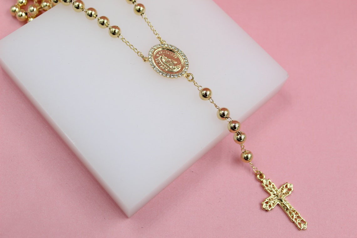 A gleaming gold rosary featuring smooth bead stations, an ornate crucifix, and a rhinestone-encrusted centerpiece rests gracefully on a white display block against a soft pink background.