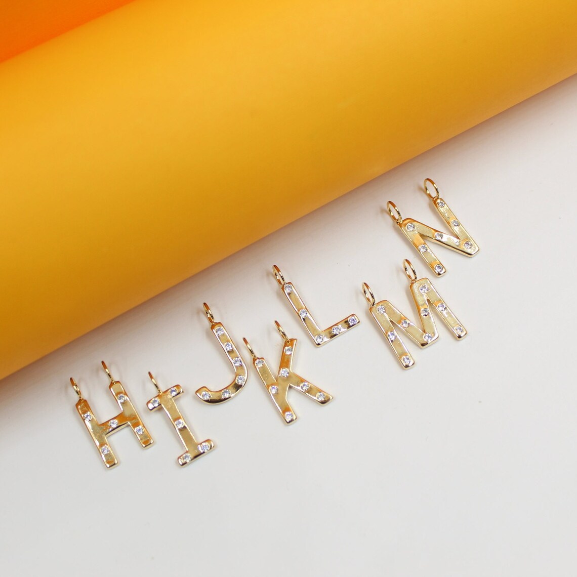 Several gold alphabet letter pendants, embellished with sparkling diamonds, are neatly arranged on a white surface against a bright orange backdrop.