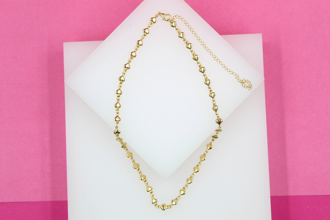 A delicate gold heart-shaped link necklace rests on a white display block against a vibrant pink background.