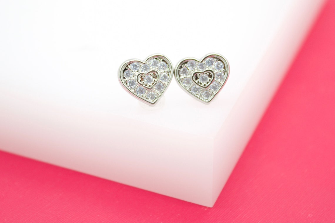 A pair of silver heart-shaped stud earrings featuring sparkling cubic zirconia crystals rests on a white display block against a vibrant pink background.