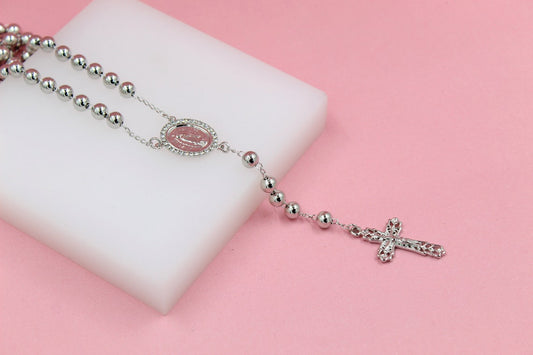 A silver rosary with shiny metallic beads rests gracefully on a white display block against a soft pink background.