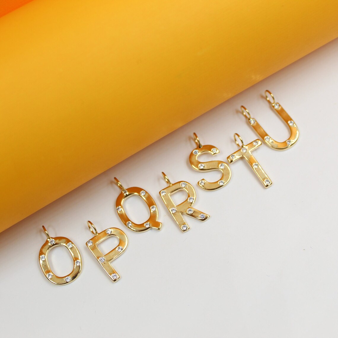 Sparkling gold alphabet letter charms O, P, Q, R, S, and T lie in a row against a vibrant orange and white background.