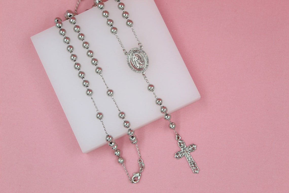 A shiny silver rosary necklace with polished ball beads rests on a white square display block against a vibrant pink background.