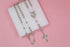 A delicate silver rosary necklace with bead chains, an oval Virgin Mary pendant, and an ornate crucifix rests on a white square display against a pink background.