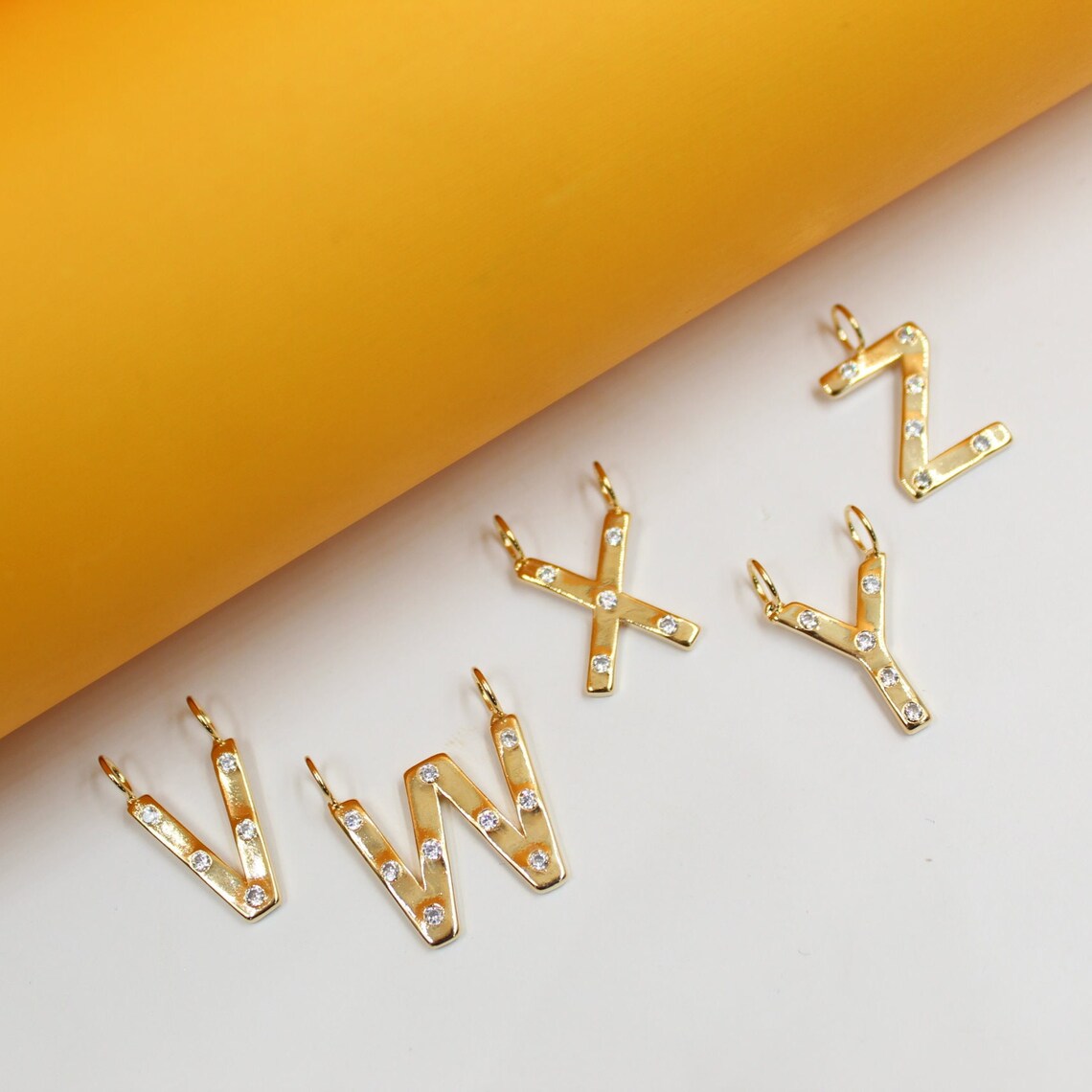 Several gold initial letter pendants, including V, W, X, Y, and Z, sparkle with embedded diamonds against a white and yellow background.