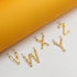 Several gold initial letter pendants, including V, W, X, Y, and Z, sparkle with embedded diamonds against a white and yellow background.