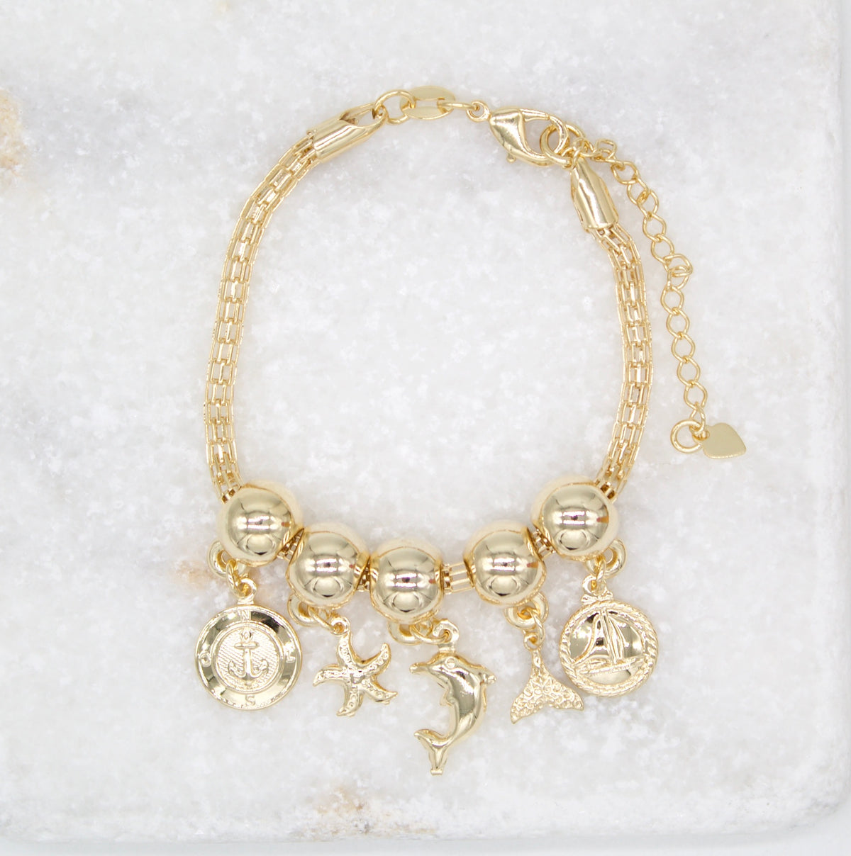 A gold charm bracelet featuring nautical-themed pendants rests on a white marble surface.
