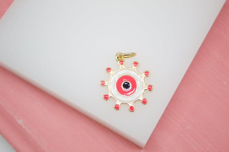A gold evil eye charm with vibrant pink enamel details rests on a white display surface against a soft pink background.