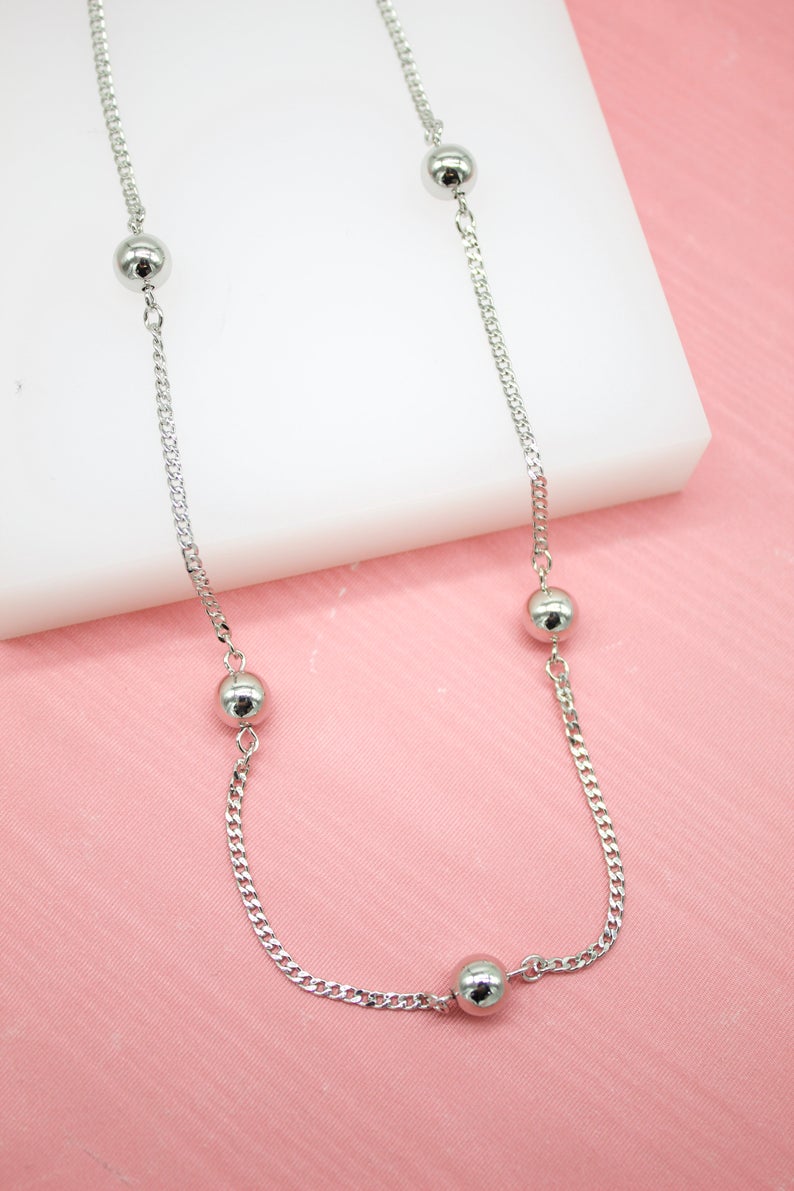 A delicate silver chain necklace features evenly spaced polished bead stations resting against a pink and white background.