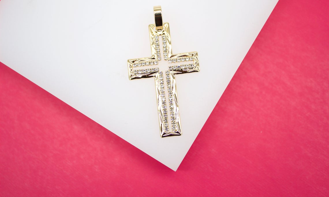 A gold diamond cross pendant rests on a white display surface against a vibrant pink background.