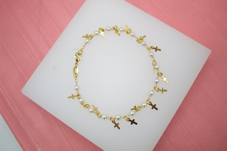 A delicate gold bracelet featuring alternating pearls and small cross charms rests on a white display surface against a pink background.