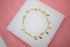 A delicate gold bracelet featuring alternating pearls and small cross charms rests on a white display surface against a pink background.