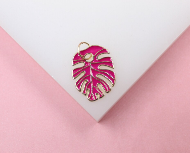 A vibrant pink enamel monstera leaf charm with gold trim rests on a white display against a pink background.