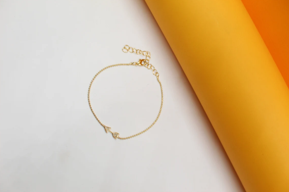 A delicate gold chain bracelet featuring two sparkling triangular diamond charms rests on a white surface next to bright yellow and orange paper rolls.