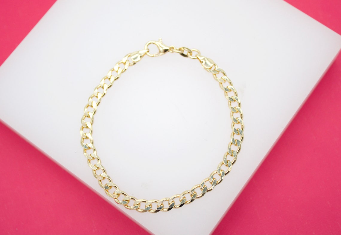 A delicate gold curb chain bracelet rests on a white display surface against a vibrant pink background.