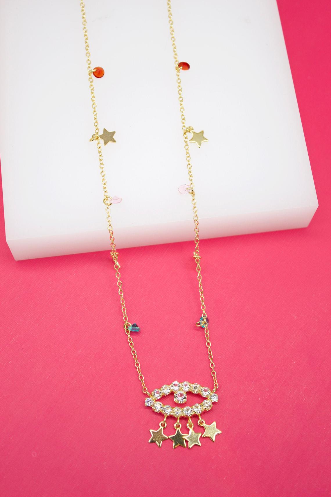 A delicate gold chain necklace drapes across a white and pink background, featuring sparkling star charms and a prominent crystal-embellished evil eye pendant.