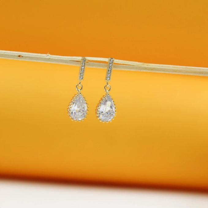 A pair of gold drop earrings featuring teardrop-shaped cubic zirconia stones hangs delicately from a wooden twig against a warm orange background.
