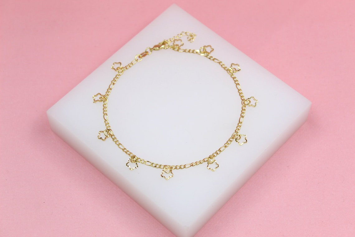 A delicate gold flower charm anklet rests on a white display block against a soft pink background.